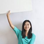 Versus Billboard - Cheerful Asian woman sitting cross legged on floor against white wall in empty apartment and showing white blank banner