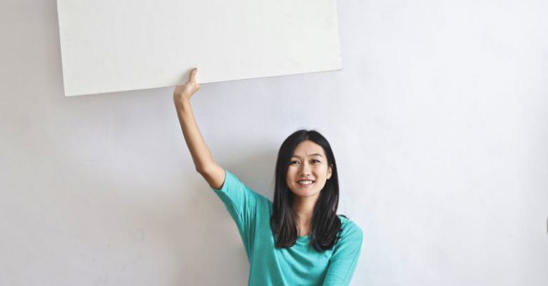Versus Billboard - Cheerful Asian woman sitting cross legged on floor against white wall in empty apartment and showing white blank banner