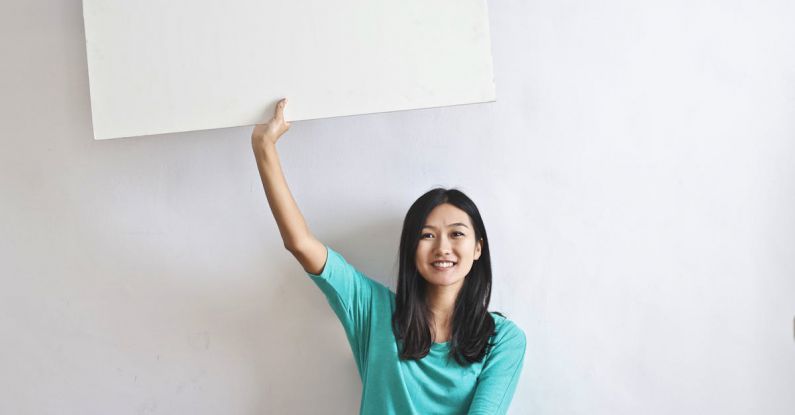 Versus Billboard - Cheerful Asian woman sitting cross legged on floor against white wall in empty apartment and showing white blank banner