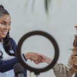 Handshake Influencer - Smiling multiethnic females wearing formal outfits shaking hands and looking at each other friendly while sitting against round video light after interview