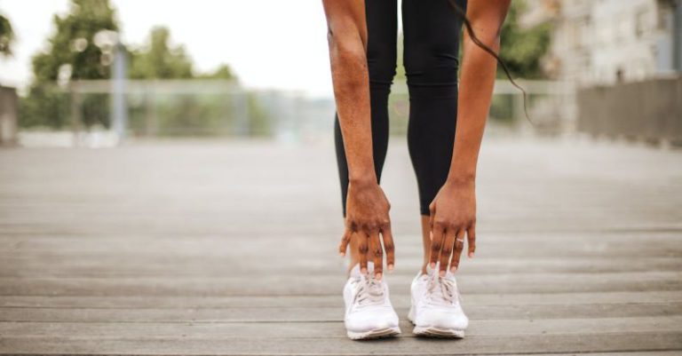 Sports Sneakers - From below crop slender female athlete in sportswear and white sneakers doing standing forward bend exercise for stretching body on wooden floor of street sports ground against blurred urban environment in daytime